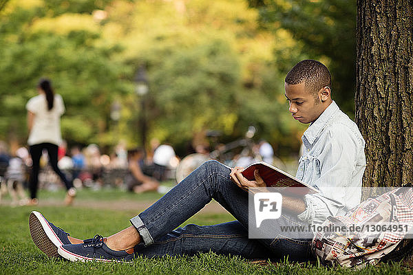 Side view of man studying while sitting by tree trunk in campus