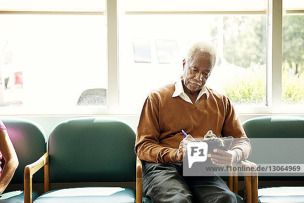 Älterer Mann schreibt auf Klemmbrett  während er auf einem Stuhl im Wartezimmer sitzt
