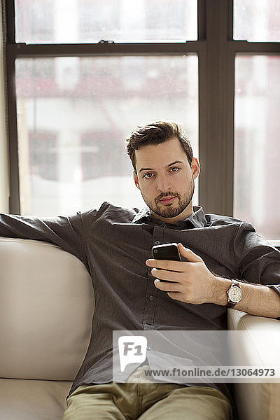Porträt eines Mannes  der ein Smartphone in der Hand hält  während er zu Hause auf dem Sofa sitzt