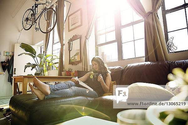 Frau liest Buch  während sie an einem sonnigen Tag zu Hause auf dem Sofa Tee trinkt