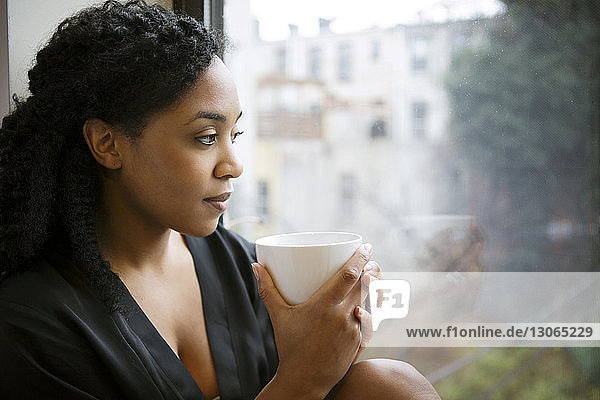 Woman with cup looking away while sitting against window