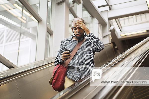 Mann mit Kopf in der Hand benutzt Smartphone  während er auf der Rolltreppe am Bahnhof steht