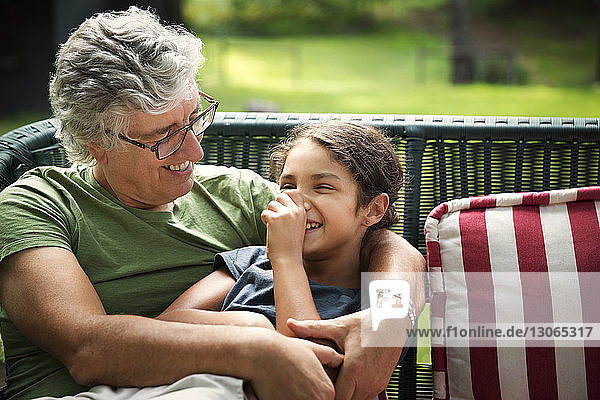 Lächelnder Großvater umarmt Enkelin  während er auf einem Korbsofa im Hof sitzt