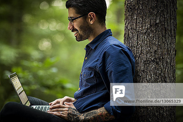 Mann benutzt Laptop-Computer  während er im Wald an einem Baum sitzt