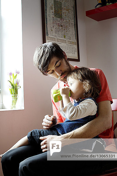 Father looking at girl drinking while sitting against window