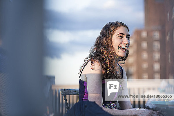 Fröhliche Frau lacht  während sie auf einem Stuhl gegen den Himmel sitzt