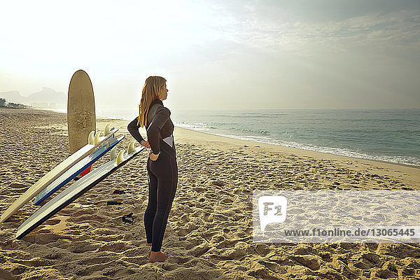 Woman putting on wetsuit while standing by surfboards at beach