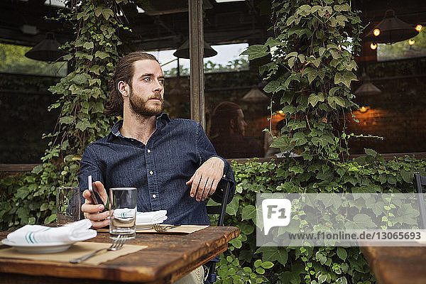 Nachdenklicher Mann hält Smartphone in der Hand  während er im Straßencafé sitzt