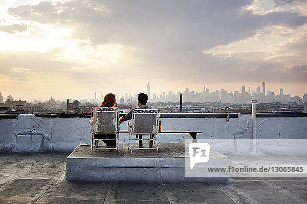 Rückansicht eines Paares  das sich bei Sonnenuntergang auf Stühlen auf der Gebäudeterrasse ausruht