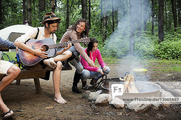 Woman roasting marshmallows while sitting by friend playing guitar at campfire in forest