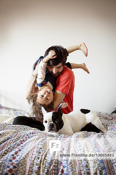 Portrait of father playing with daughter and dog while sitting on bed against wall