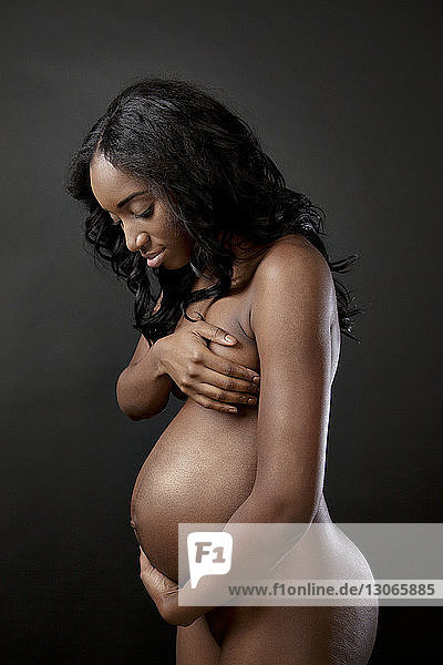 Seitenansicht einer schwangeren Frau  die die Brust bedeckt  während sie vor schwarzem Hintergrund steht