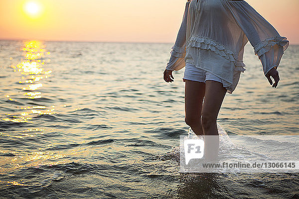 Mittelteil einer Frau  die bei Sonnenuntergang am Strand im Wasser geht