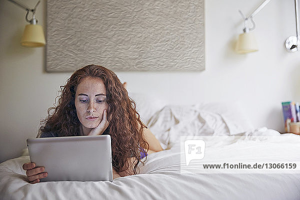 Frau schaut auf Tablet-Computer  während sie im Bett an der Wand liegt