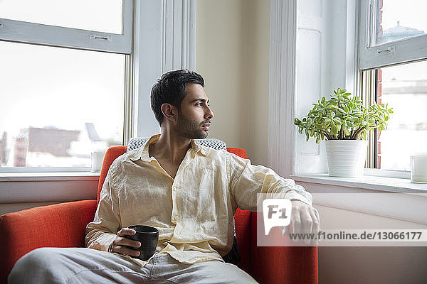 Mann mit Kaffeetasse schaut weg  während er zu Hause auf einem Sessel sitzt