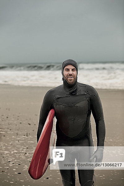 Porträt eines glücklichen Mannes  der ein Surfbrett trägt  während er am Strand steht