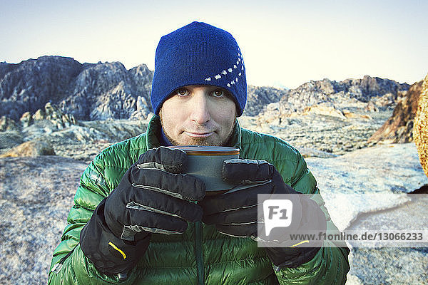 Portrait of man holding coffee cup while sitting on rocks during winter