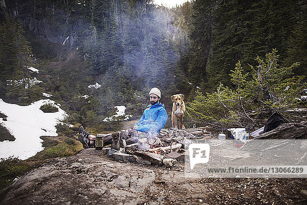 Mann sitzt mit Hund am Lagerfeuer im Wald