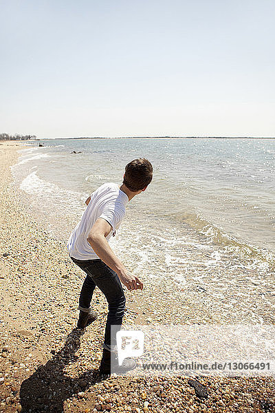Rückansicht eines Mannes  der am Ufer stehend Steine ins Meer wirft