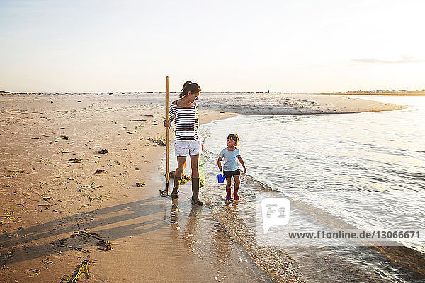 Mother and daughter walking with rake on seashore during sunny day
