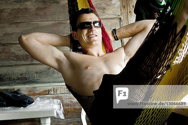 Smiling shirtless man wearing sunglasses resting in hammock at log cabin