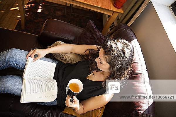 Hochwinkelansicht einer Frau  die ein Buch liest  während sie an einem sonnigen Tag zu Hause auf dem Sofa Tee trinkt