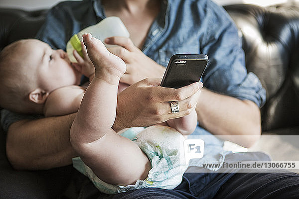 Mittelsektion eines Mannes  der seinen Sohn füttert  während er zu Hause ein Mobiltelefon benutzt