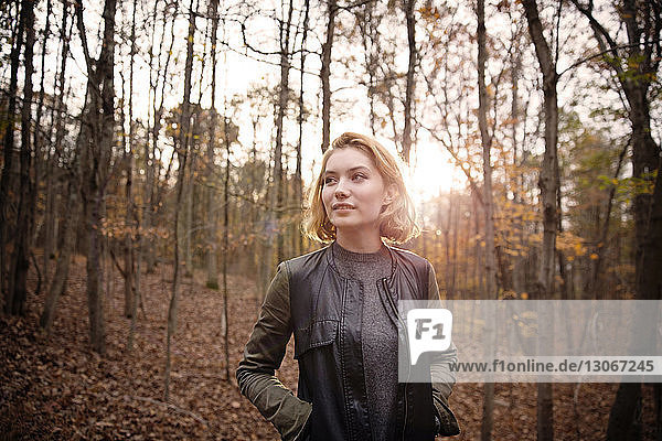 Thoughtful woman with hands in pockets standing against trees in forest during winter