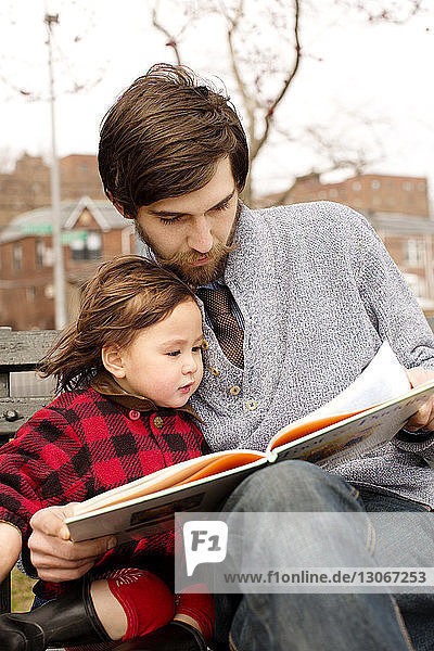 Vater und Tochter schauen sich ein Buch an  während sie im Park auf einer Bank sitzen