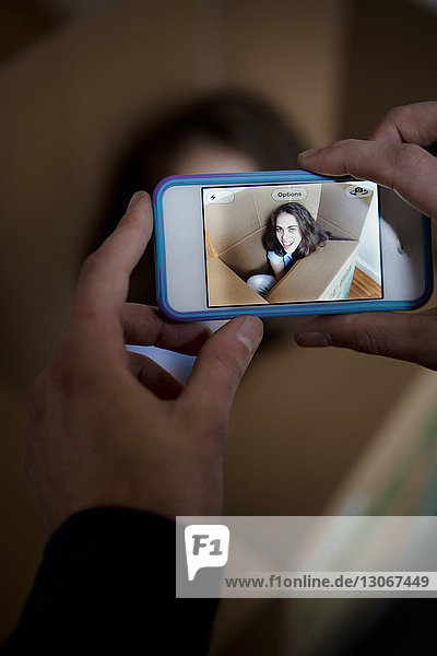 Ausschnitt eines Mannes  der eine Frau fotografiert  die zu Hause in einem Pappkarton sitzt