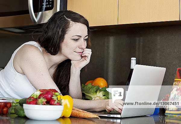 Frau benutzt Laptop-Computer  während sie sich zu Hause an die Küchentheke lehnt