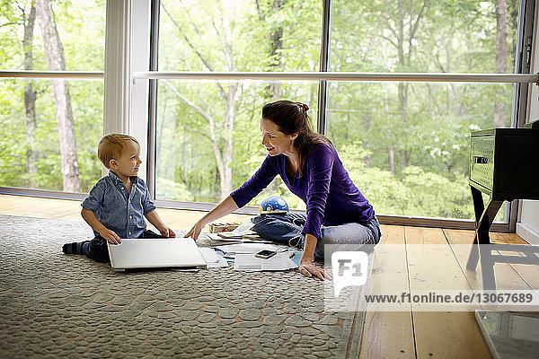 Mutter macht Papierkram und sieht ihren Sohn mit Laptop-Computer am Fenster am Boden sitzen