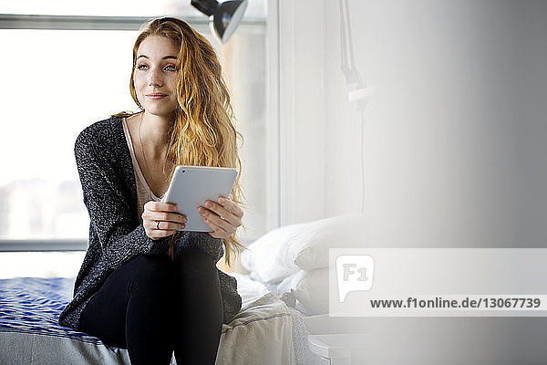 Frau mit Tablet-Computer schaut weg  während sie zu Hause auf dem Bett sitzt