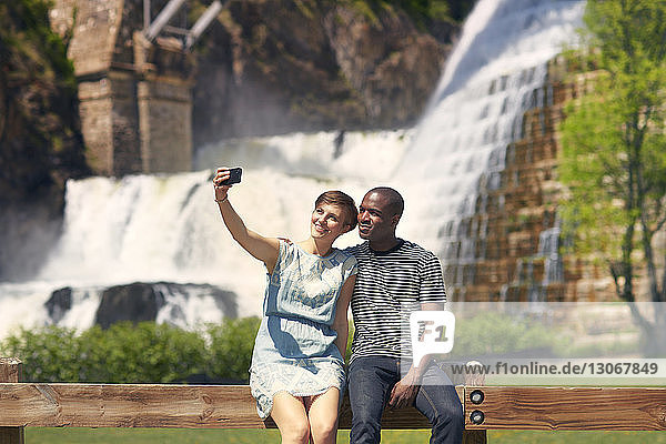 Couple taking selfie while sitting on railing against waterfall
