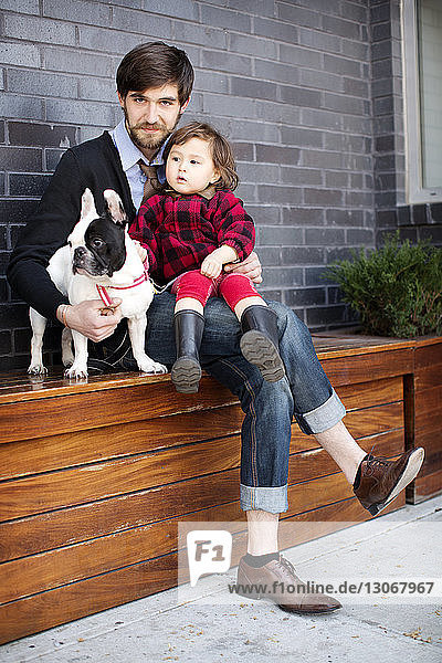 Porträt von Vater und Tochter mit Hund auf Bank an der Wand sitzend