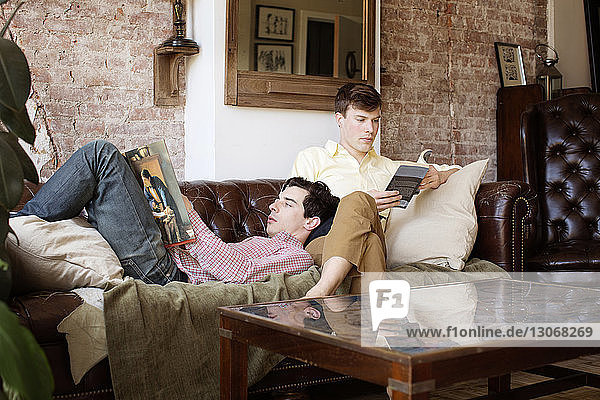 Schwules Paar liest Bücher  während es sich zu Hause auf dem Sofa entspannt
