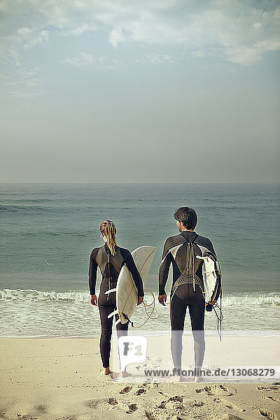 Rear view of couple carrying surfboards while standing at shore