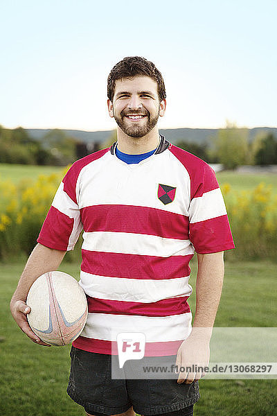 Porträt eines glücklichen Spielers  der den Rugbyball auf dem Spielfeld gegen den klaren Himmel hält