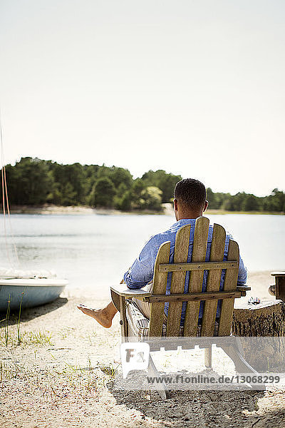 Rear view of man relaxing on adirondack chair at lakeshore