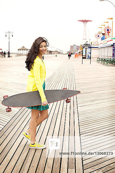 Porträt einer Teenagerin  die ein Skateboard trägt  während sie auf dem Bodenbrett gegen den klaren Himmel läuft