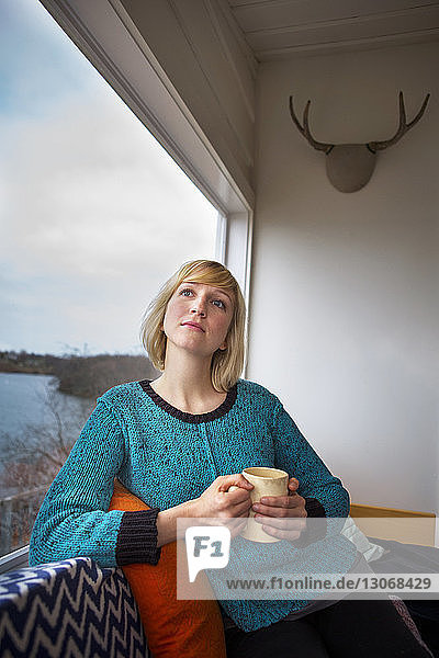 Nachdenkliche Frau trinkt Kaffee  während sie zu Hause durchs Fenster schaut