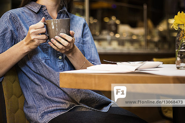 Mittelteil einer Frau mit Kaffeetasse in der Hand  die in einem Café am Tisch sitzt