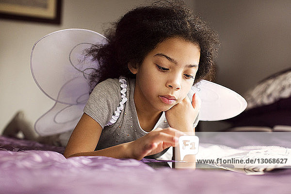 Mädchen in Feenflügeln benutzt Tablet-Computer  während sie zu Hause auf dem Bett liegt