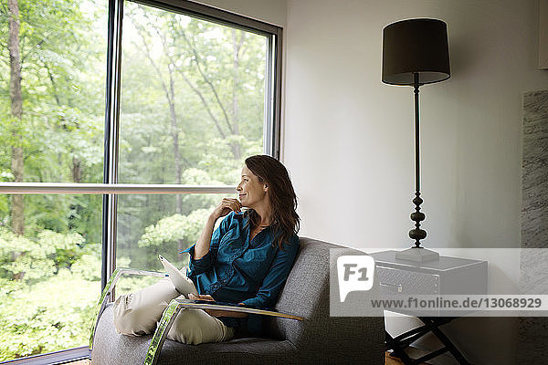 Nachdenklich lächelnde Frau mit Tablet-Computer auf dem Sofa sitzend  während sie zu Hause durchs Fenster schaut