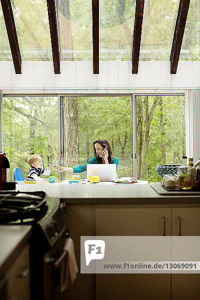 Glückliche Mutter mit Technologie  die ihrem Sohn an der Küchentheke Essen gibt