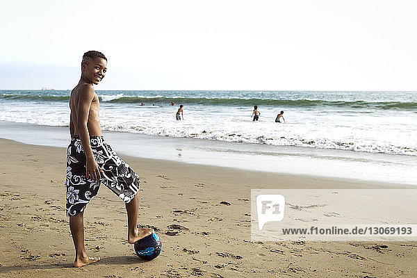 Porträt eines Jungen  der mit einem Bein auf einem Fussball am Strand steht