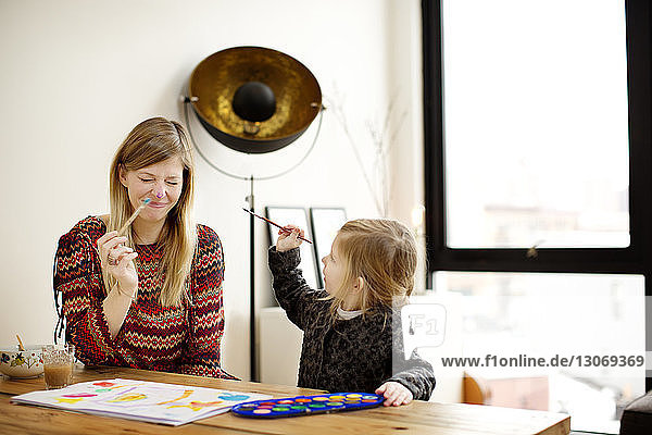 Playful mother and daughter painting while sitting at table in home