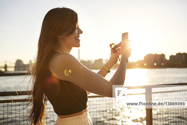 Happy woman photographing with mobile phone by river during sunset