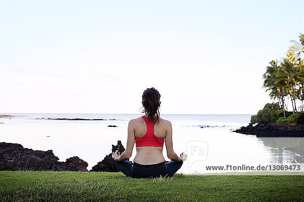 Rear view of woman meditating while sitting on field against sea