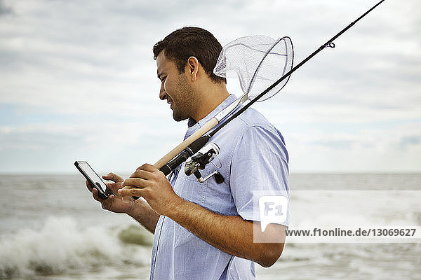 Man holding fishing rod using mobile phone while standing by sea
